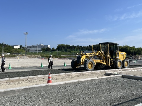 ヘルメットを被った作業員が見守る中、黄色のボディの工事車両がトラック部分の路盤工事を行っている様子の写真