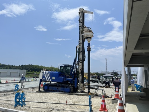 トラックと観覧スタンドの間で、青色の工事車両が円柱の杭を掴み地面に垂直にあてている、写真判定室棟基礎部分の杭打設工事の様子写真