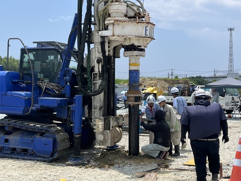 青色の工事車両が円柱の杭を掴み地面に垂直に埋め込み、ヘルメットを被った作業員の方々が杭の長さを計測している、写真判定室棟基礎部分の杭打設工事の様子写真