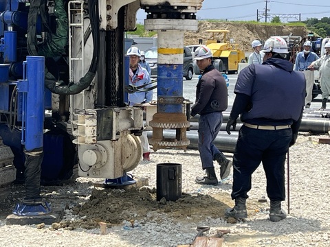 円柱の杭を地上から約50センチメートルくらい出して地面に埋め込んだ青色の工事車両が杭を離した様子を、ヘルメットを被った作業員の方々が見ている写真判定室棟基礎部分の杭打設工事の様子写真