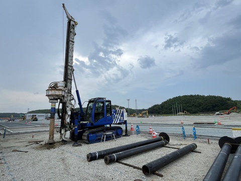 円柱の杭が地面に並んで置かれている左側で、青色の工事車両が杭を地面に埋め込んでいる写真判定室棟基礎部分の杭打設工事の様子写真