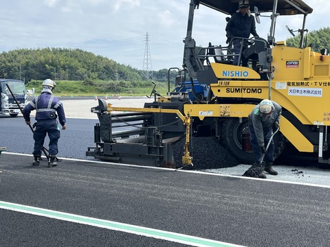 黄色のアスファルトフィニッシャーから出たアスファルトを作業員が路盤の上に敷均している、トラック部分の基層工事の様子をアップで撮影した写真
