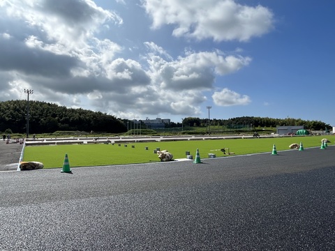 トラックとフィールドの境目に緑色の三角コーンが一列に並び、広大なフィールド部分に手前から人工芝の敷設工事が行われている様子の写真