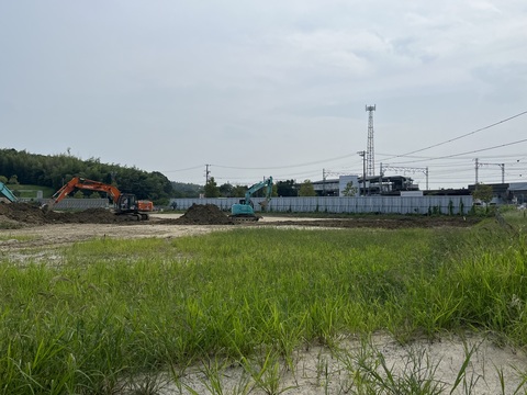雑草が生えている交流広場の工事予定地で、水色のショベルカーとオレンジ色のショベルカー2台が基盤造成工事を行っている写真