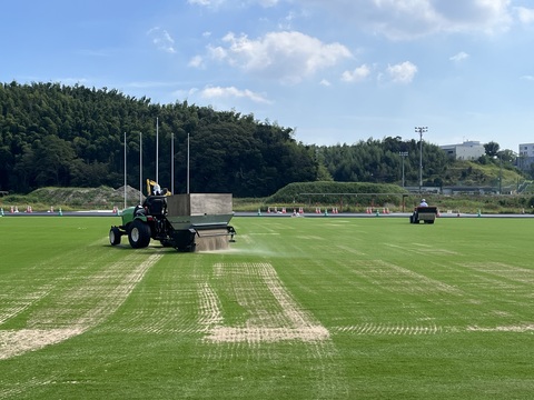 人工芝を敷き詰めた広大なフィールドで、後部にローラーが付いている赤色の工事車両と、砂を撒いている緑色の工事車両が砂を敷設している工事の様子をアップで撮影した写真