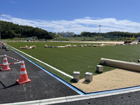 広大なフィールド内に人工芝が敷き詰められ、中央部分に人工芝を包んでいたベージュ色のシートと緑色のロープが無造作に置かれている敷設工事の様子写真