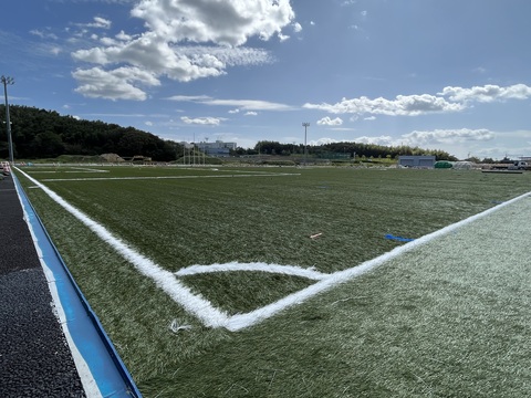 フィールド部分に人工芝が敷き詰められ、ラインが引かれたサッカーコートの角から敷設工事の様子を撮影した写真