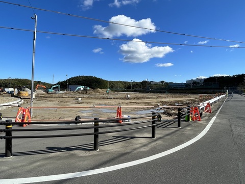 道路の境目の工事用鋼板塀が撤去された工事予定地の交流広場の奥の砂が山積みされた場所で、工事車両が作業を行っている様子の写真