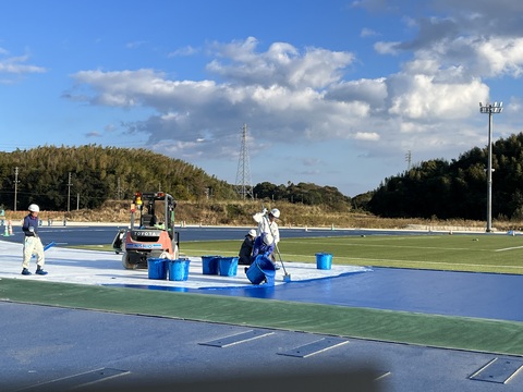 トラックの左半分に敷かれた白いシートの上にヘルメットを被った作業員が乗り、塗装が入った大きな水色のバケツをトラック部分に傾けているベース層工事の様子写真