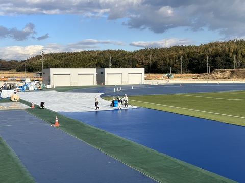 トラックに敷かれた白いシートの上にヘルメットを被った複数の作業員が乗り、ベース層工事が行われている様子を観覧スタンドから撮影した写真