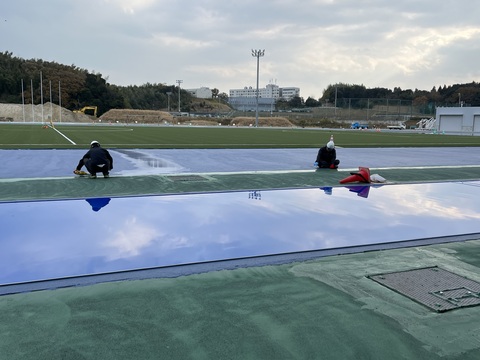アウトフィールド部分に間隔を開けて座っている2名の作業員が、トラック部分の耐久層の舗装工事を行っている様子の写真