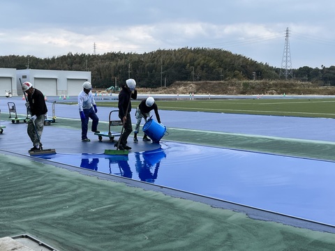 作業員が青色の塗装が入った大きな水色のバケツをトラック部分に傾けて流し、ヘルメットを被った2名の作業員が道具を使用し均している、耐久層の舗装工事を行っている写真