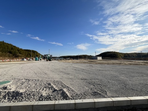 交流広場の工事予定地の奥で工事車両が地面を砕石や砂利で敷きならしている、路盤工事の様子写真