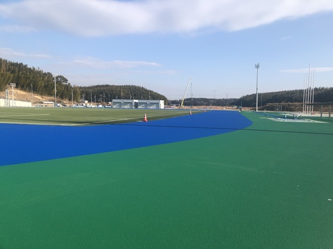 奥に器具庫が建っているのが見える、右側にカーブしたトラック部分に青色の舗装工事が行われた様子の写真
