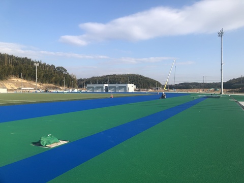 奥に器具庫が建っているのが見える、真っすぐに奥まで延びたトラック部分で青色の舗装工事が行われている様子の写真