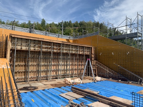 床に脚立などの道具や木材などが置かれた陸上競技場観覧スタンド部分の配筋工事と型枠設置工事が行われている様子を左斜めから撮影した写真
