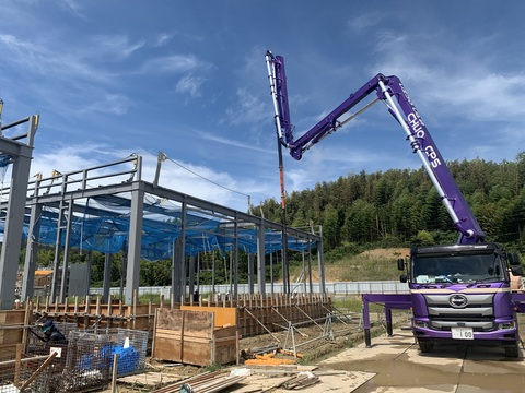 鉄骨が組まれた陸上競技場器具庫腰壁部分に、長く延びたブームで紫色のボディのコンクリートポンプ車がコンクリート打設工事を行っている様子の写真