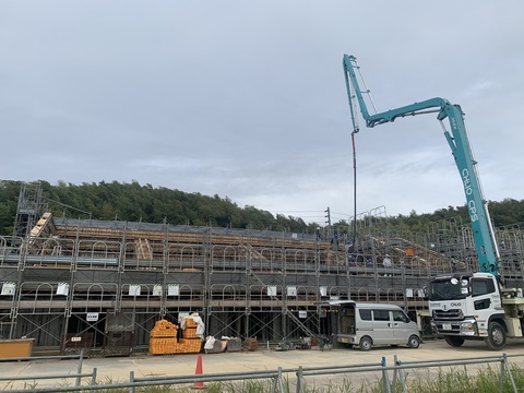 建設現場の前に資材が置かれ、軽バンとコンクリートポンプ車が停車し、延びてきたブームを使い複数の作業員が陸上競技場観覧スタンド2階部分でコンクリート打設工事を行っている様子の写真