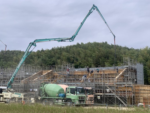 陸上競技場の建設現場の前に緑色と薄いピンク色の車体のミキサー車が並列で停車し、後方に停めたコンクリートポンプ車から長く延びたブームで陸上競技場観覧スタンド2階部分の コンクリート打設工事が行われている様子の写真