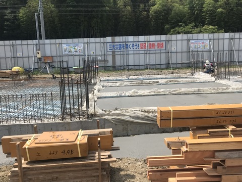 陸上競技場観覧スタンド1階床部分でヘルメットを被った作業員が枠にビニールを設置しているコンクリート打設工事が行われている手前に、場所ごとに資材が積まれている作業現場の写真