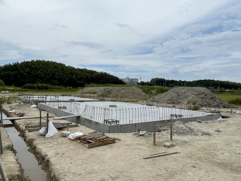 土が山積みにされた手前の陸上競技場器具庫の建設現場で、床部分のコンクリート打設工事が行われている様子の写真