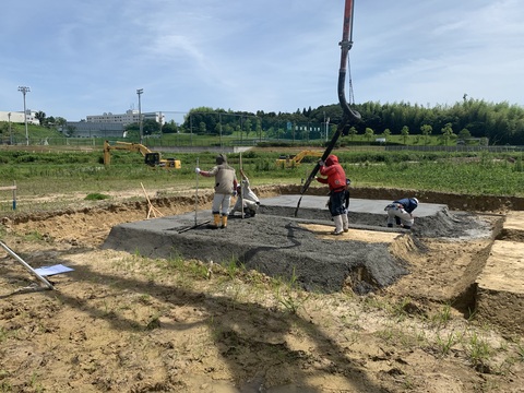 陸上競技場器具庫の建設予定地の中央部分をへこませて段差を作った地面に、4名の作業員がコンクリートの打設工事を行なっている様子の写真