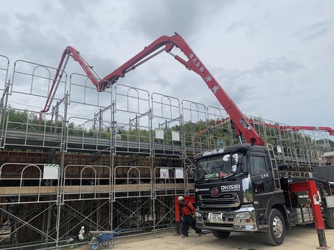 周囲に足場が組まれた陸上競技場観覧スタンドの建設現場で、コンクリートポンプ車から高所の位置まで赤色のブームが延びコンクリート打設工事が行われている写真