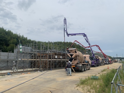 足場が組まれた陸上競技場観覧スタンドの建設現場の傍に交互に並んだ紫色や赤色のコンクリートポンプ車やミキサー車がコンクリート打設工事を行っている様子の写真