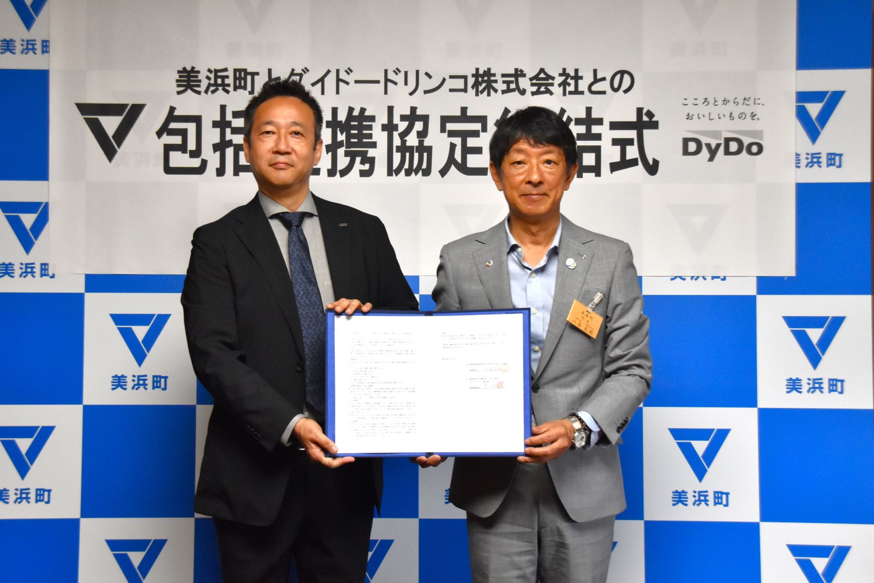 「美浜町とダイドードリンコ株式会社との包括連携協定締結式」と書かれた美浜町のバックボードの前で、スーツ姿の市長と男性が調印書を持って記念撮影している写真