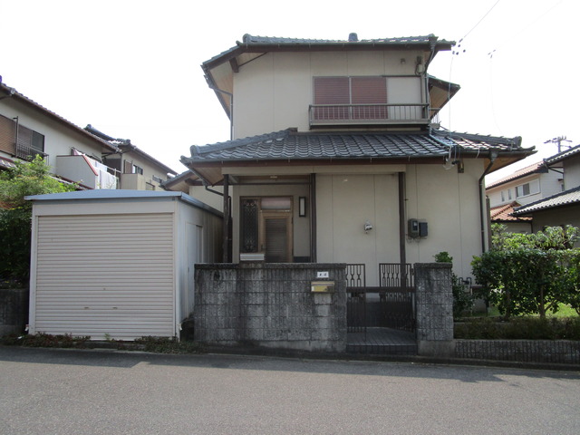 左側にシャッタ―が閉まっている駐車場があり、瓦屋根の白を基調とした2階建ての外観写真