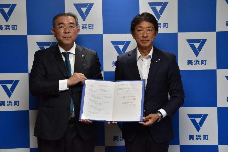 美浜町のバックボードの前に立ち、細井会長と八谷町長が一緒に協定書を持っている写真