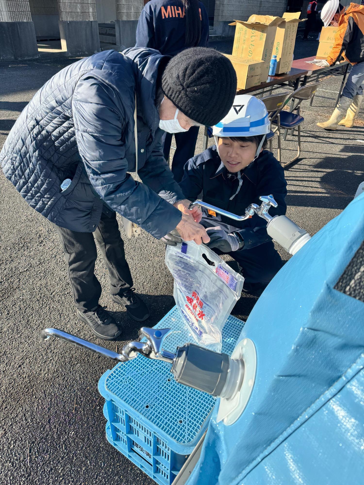 ヘルメットを被った職員と住民が簡易給水槽に付いた蛇口から給水袋へ水を給水している写真