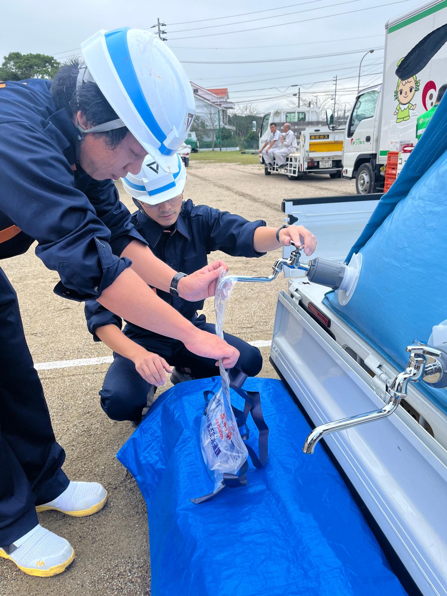 ヘルメットを被った職員の男性2名が簡易給水槽に付いた蛇口から給水袋へ水を給水している写真