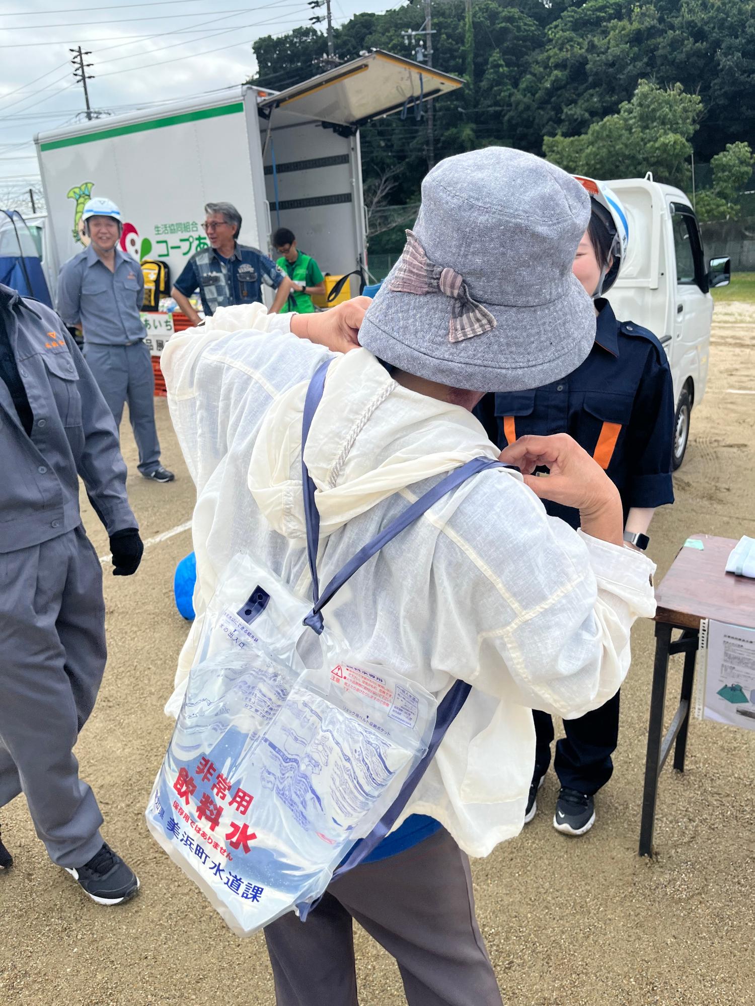 グレーの帽子を被った女性が透明な袋に水が入っている背負い式給水袋を背負い、給水袋の肩紐部分を整えようとしている様子の後ろ姿の写真