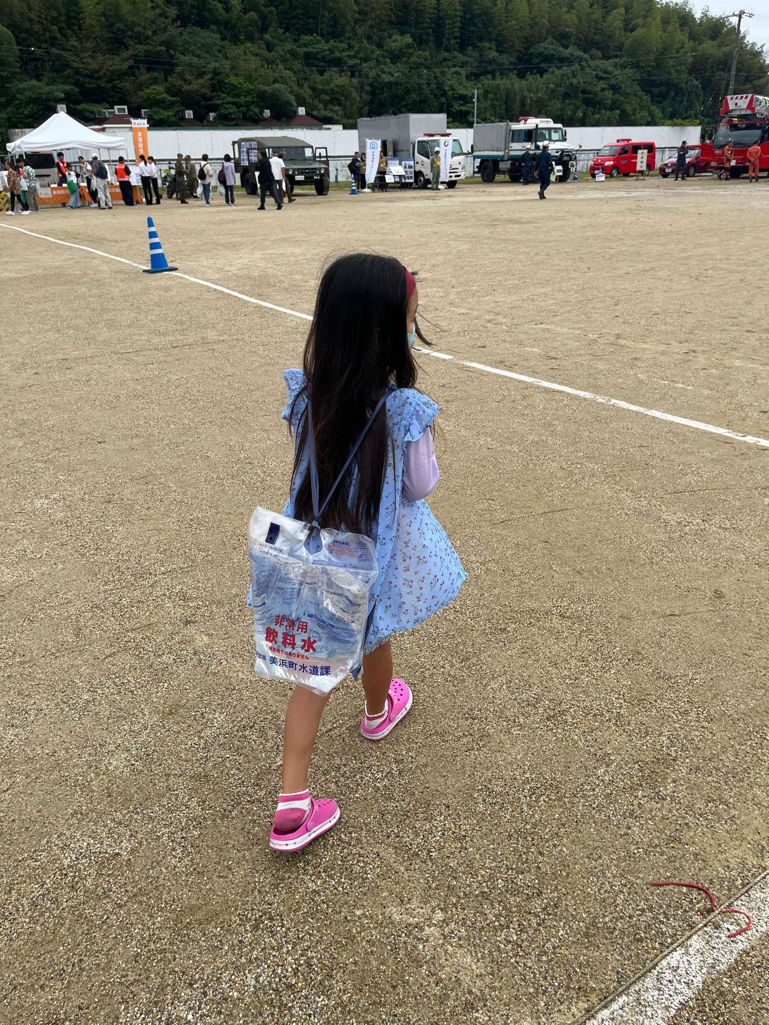 水色のワンピースを着た女の子が、水が入った「非常用飲料水」と書かれている背負い式給水袋を背負って歩いている後ろ姿の写真