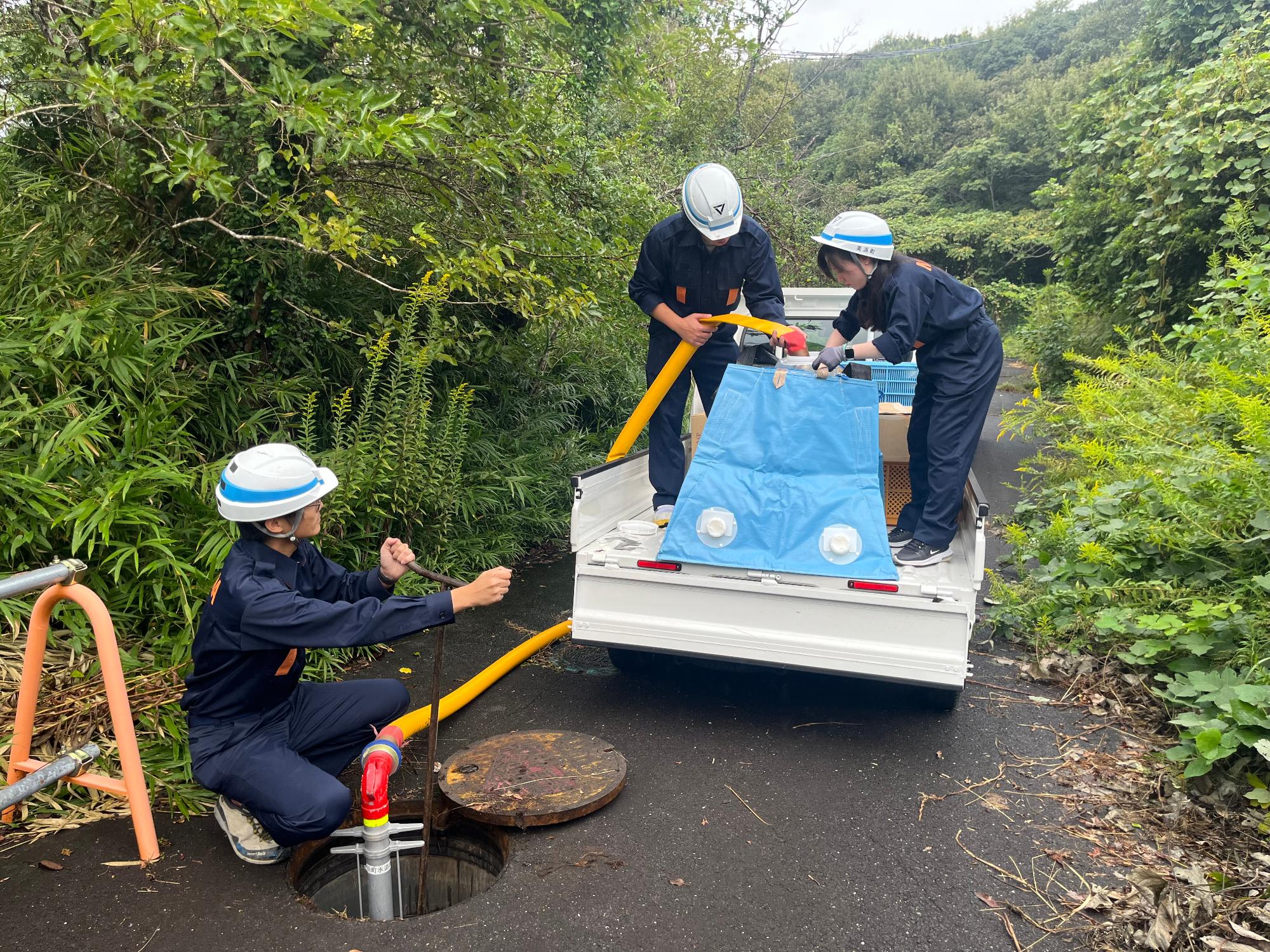 マンホールに管のような物を入れ、管の先に付いた黄色いホースを水色の簡易給水槽に繋ぎ給水を行っている様子の写真