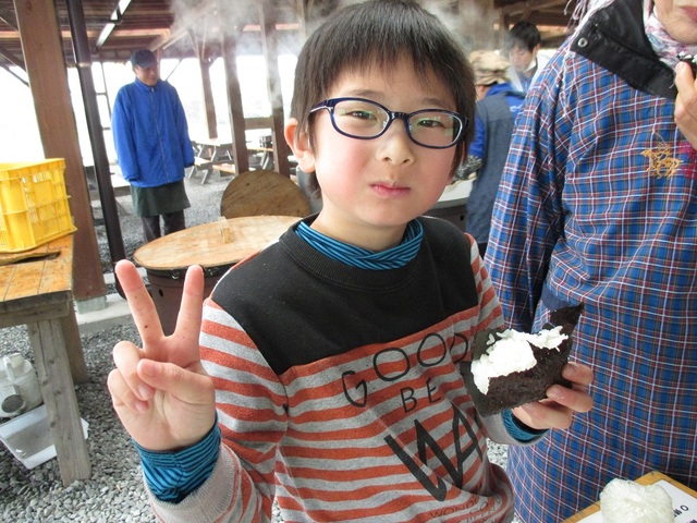 海苔に巻かれたおにぎりを左手で持ち、カメラに向かってピースサインをしている男の子の写真
