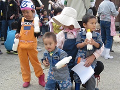 ペッボトルに入ったバターを両手に持ちポーズをとるオレンジの服を着た男の子としゃがんでいる女性に支えられている2人の男の子の写真