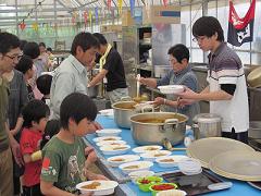 カレーを容器によそっている方々と、テーブルに並べられたカレーを取っている子供たちの写真