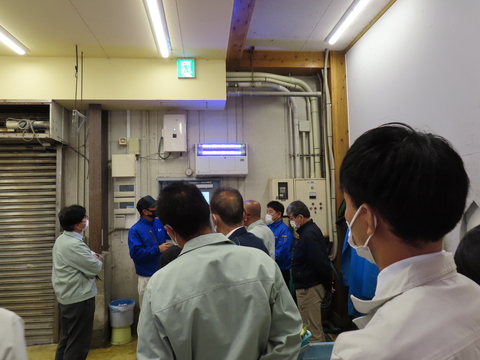 現地見学で参加者たちが集まり、前方の方を向いて説明を聞いている様子を後方から写した写真