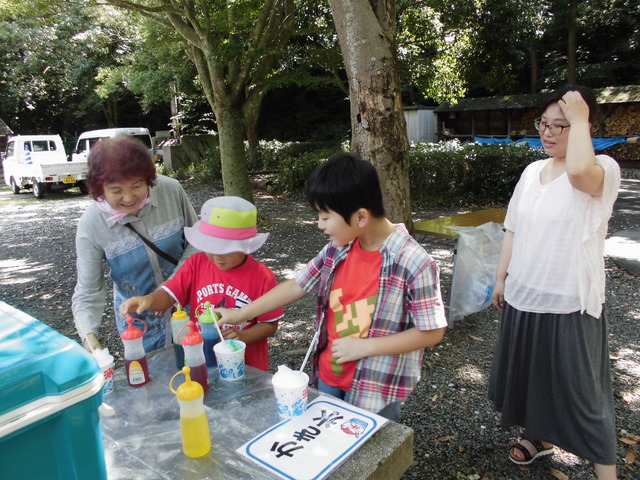 屋外の木陰の下に設置されている「かき氷」と書かれた紙が置かれた台で、子供たちが氷にかけるシロップを選んでいる写真