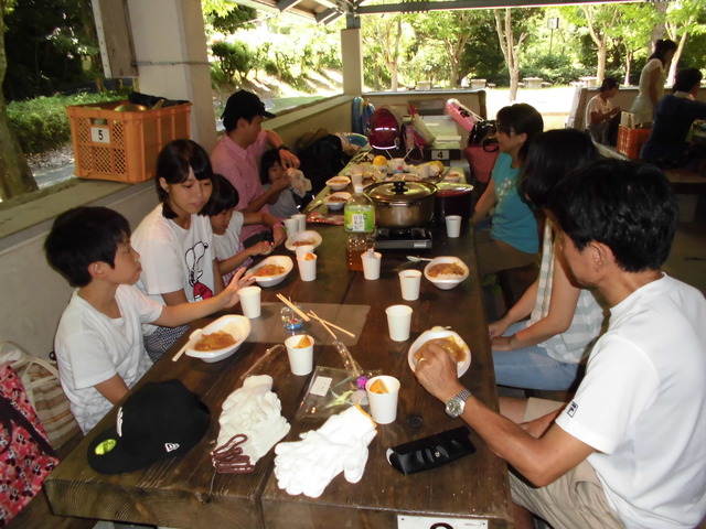 完成したカレーが長いテーブルに並べられ、参加者たちが向かい合って座っている写真