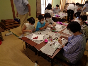 座卓タイプの長机が設置された和室で、参加者たちが繭を使ってクラフト作りを行っている写真