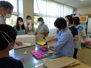 参加者たちが作業台の周りに集まり、ストライプ柄のシャツを着た女性が鍋に入った黄色い液体状の石鹸を型に流し込んでいる写真