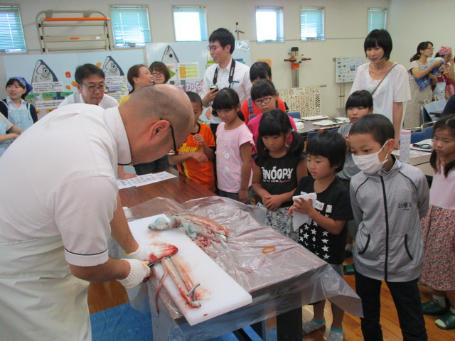 男性がまな板の上でウナギをさばいており、子ども達が集まってさばいている様子を興味深く見学している写真