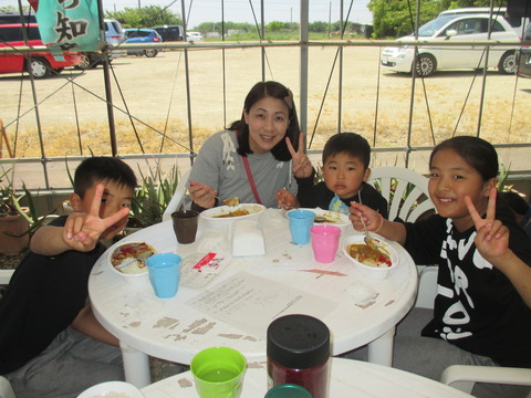 男の子2人、お母さん、女の子が丸いテーブルに座ってカレーを食べており、カメラに向かってピースサインをしている昼食の様子の写真