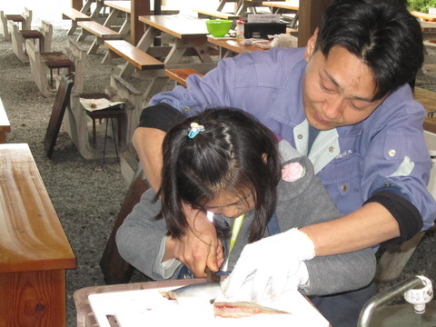 まな板の上に置かれた魚を真剣な様子でさばいている女の子と、女の子の後ろに立って手を添えて一緒に手伝っている関係者の写真