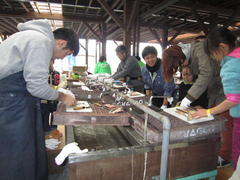 奥に続く流し台を両側から向かい合う形で立ち並んでいる参加者達が、まな板の上に置かれた魚を調理している様子の写真