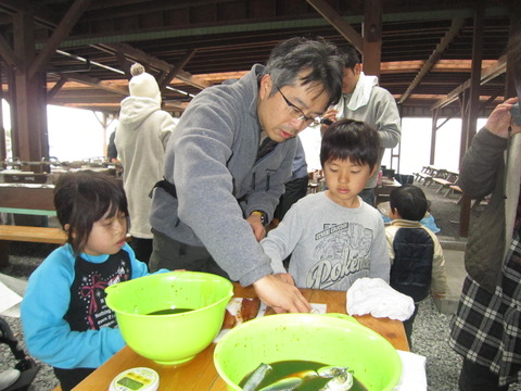 緑色のボウルの中に調味液に漬けられた魚、まな板の上に捌かれた魚の切り身が並べられ、両側に立っている子ども達に教えるように作業をしている男性の写真