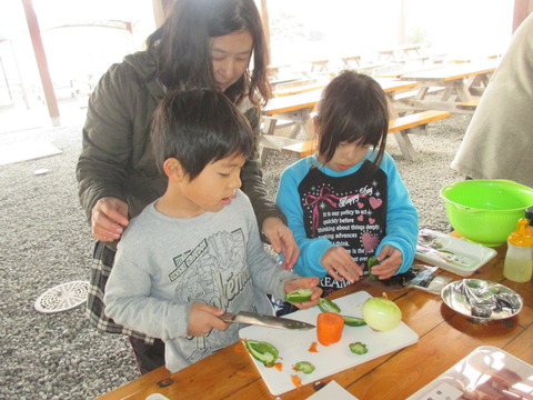 様々な食材が用意された調理台の上にまな板が置かれ、2人の子どもが関係者に見守られながらニンジンやピーマン、玉ねぎなどの食材をカットしている様子の写真